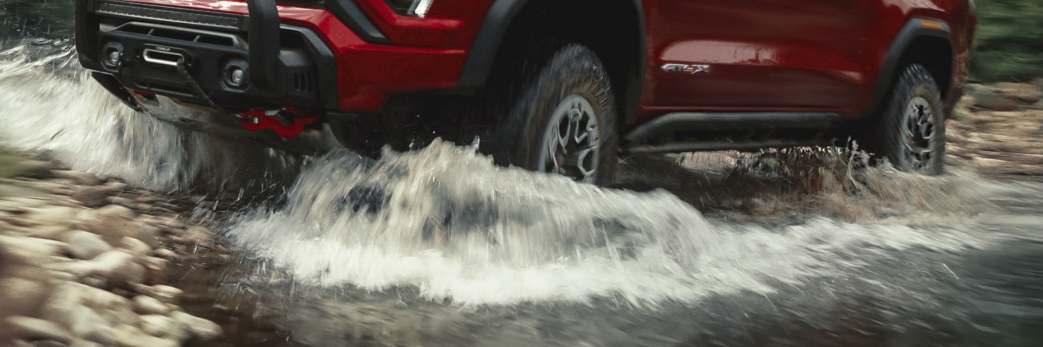 A Red 2027 GMC Canyon Driving Through Shallow Water Causing Splashes