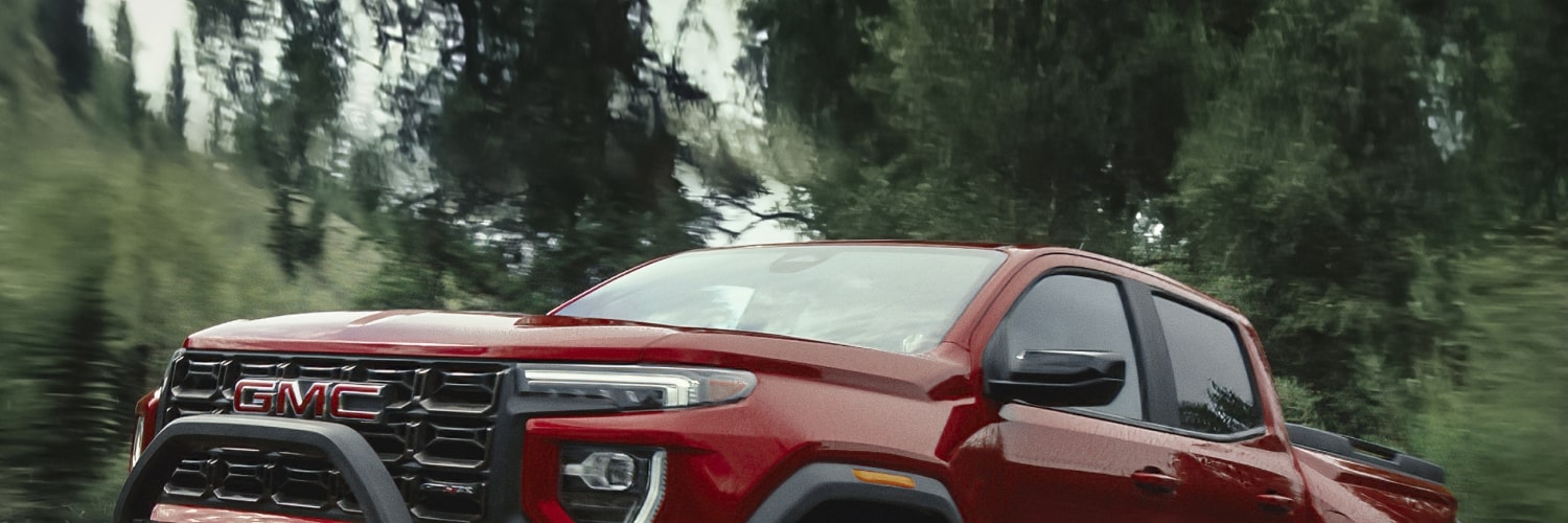 A Red 2027 GMC Canyon Driving Through Shallow Water in a Lush Green Environment