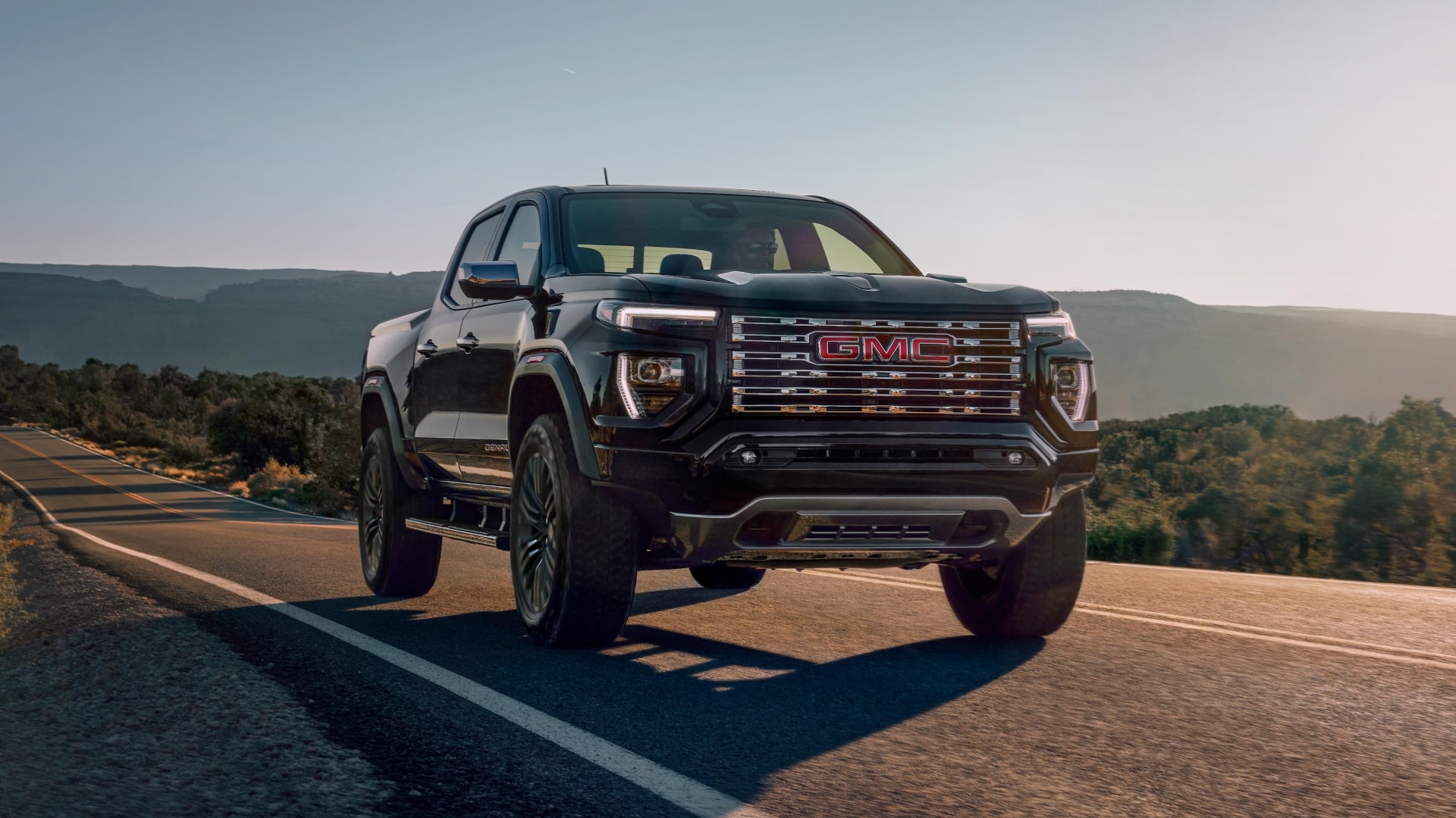 Front View Of a Black 2027 GMC Canyon Denali Truck on a Paved Road With Mountains in the Background
