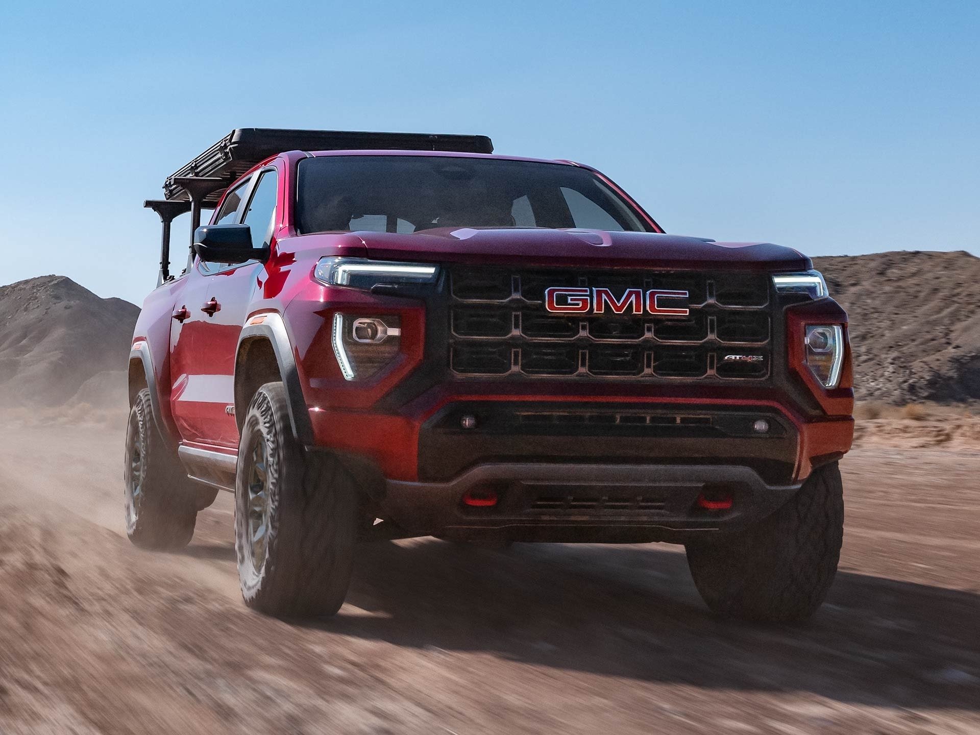 A Red 2027 GMC Canyon AT4X with a Roof Rack Driving on a Dusty Off-Road Trail Under a Blue Sky