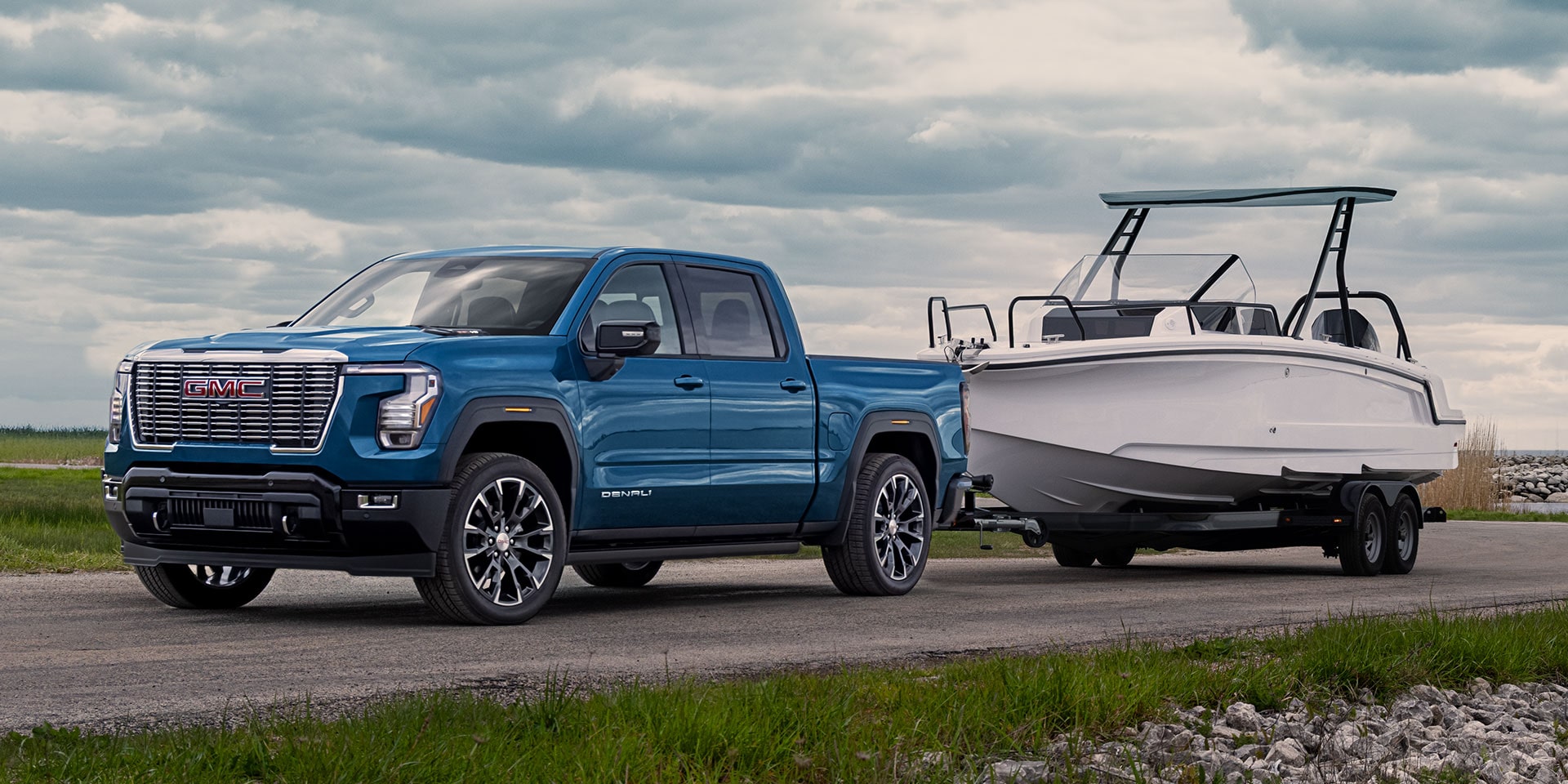 A Blue 2027 GMC Sierra 1500 Denali Truck Towing a White Boat on a Road under a Cloudy Sky