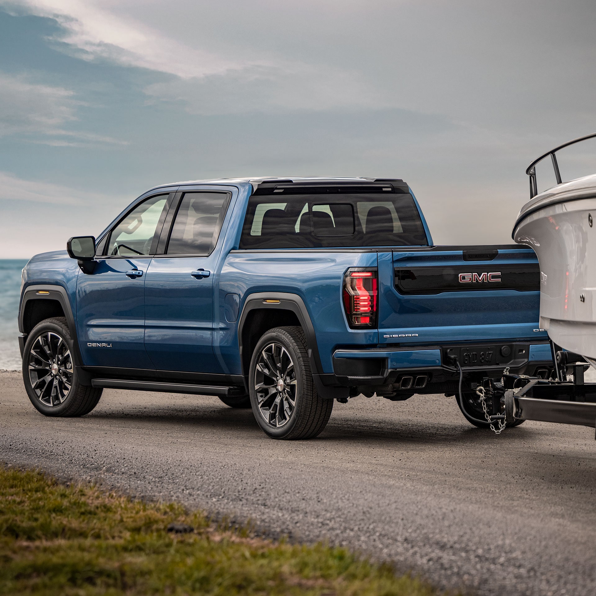 A Rear 3/4 View of a Blue GMC Sierra 1500 Denali Towing a White Boat by a Body of Water