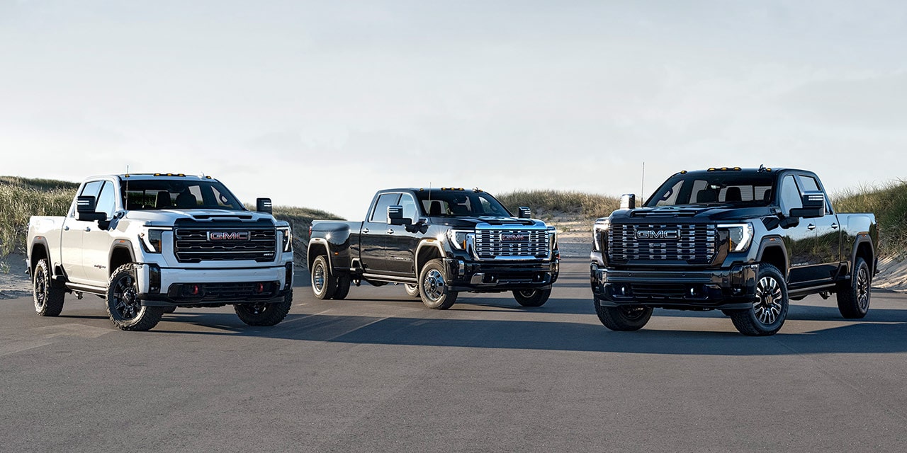 A White GMC Sierra HD AT4, a Dark Gray GMC Sierra HD Denali, and a Black GMC Sierra HD Denali Ultimate on a Road with Sand Dunes