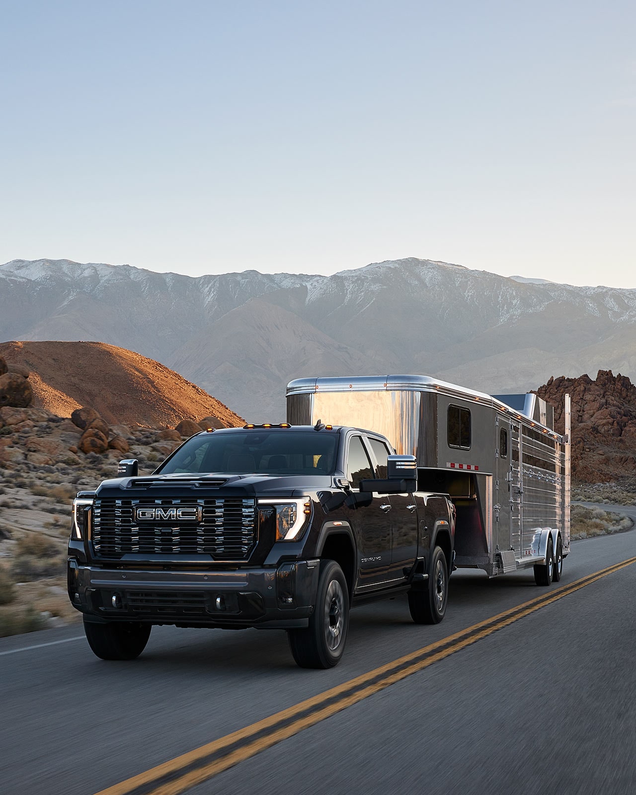 A Black 2027 GMC Sierra HD Truck Drives On a Paved Road, Towing a Silver Horse Trailer in a Rocky Desert Landscape with Mountains in the Background