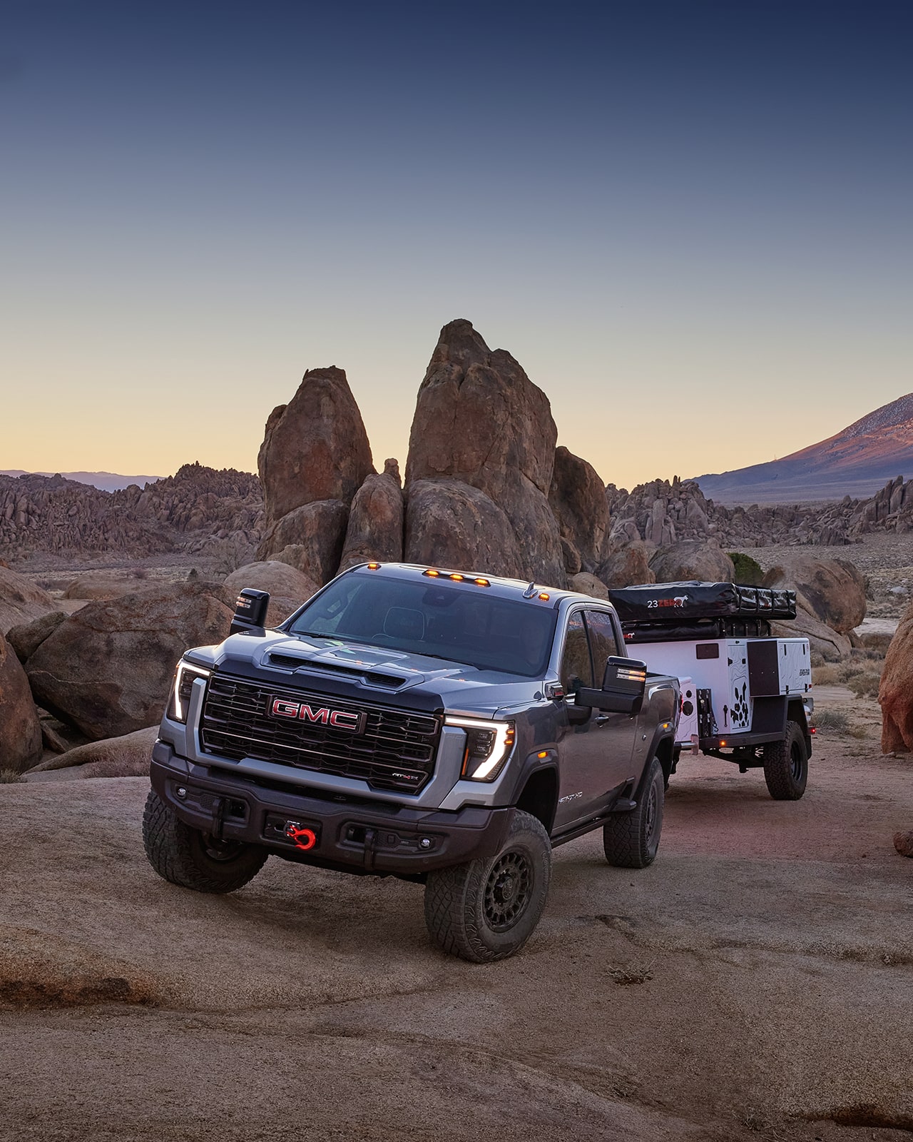 A Dark Gray 2027 GMC Sierra HD Truck with Red Recovery Hooks and Off-Road Tires Is Parked On a Rocky Desert Landscape, Towing a Small White Adventure Trailer