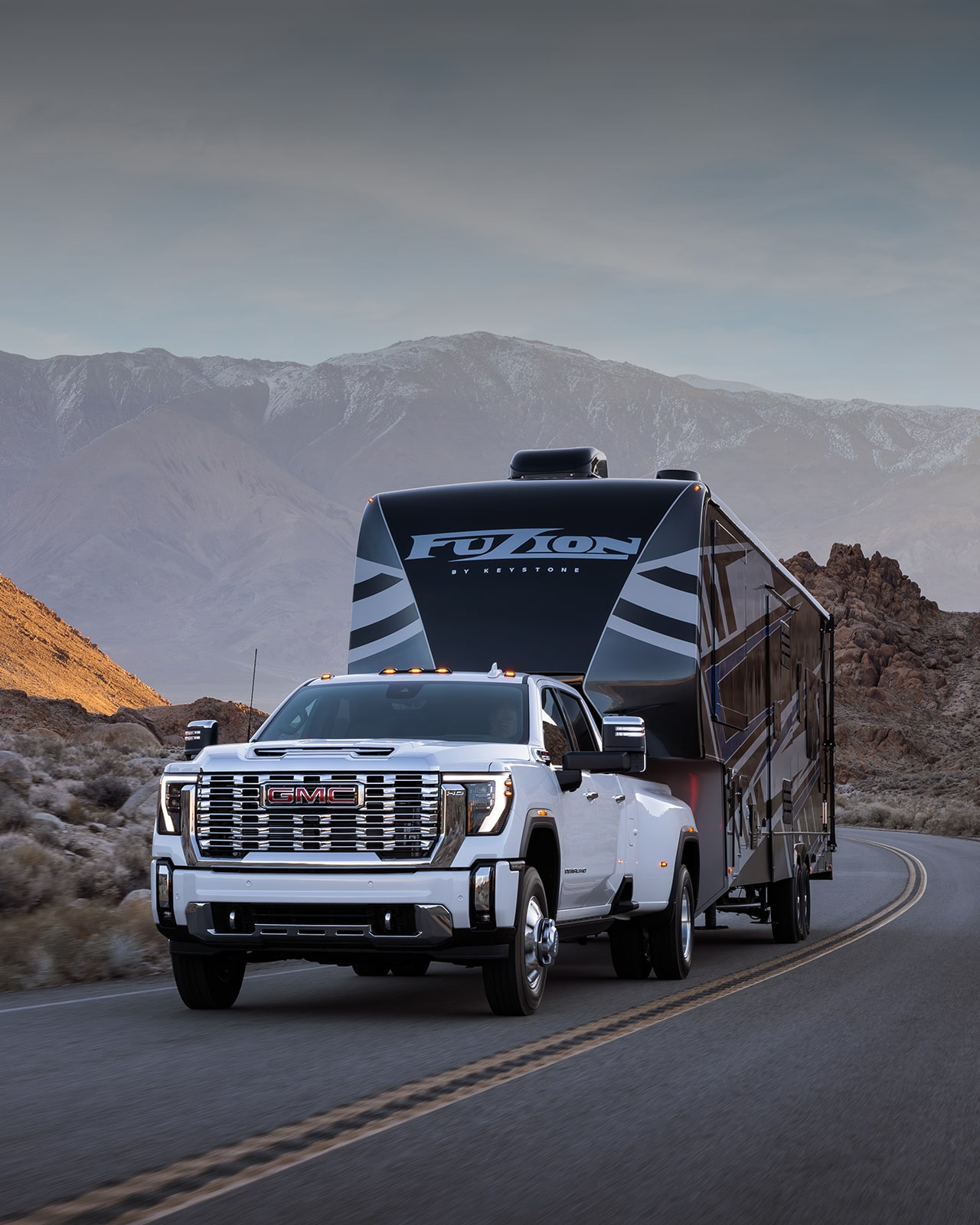 A White 2027 GMC Sierra HD Dually Truck Drives On a Paved Road Towing a Large Black and White Fifth-Wheel RV in a Desert Mountain Setting