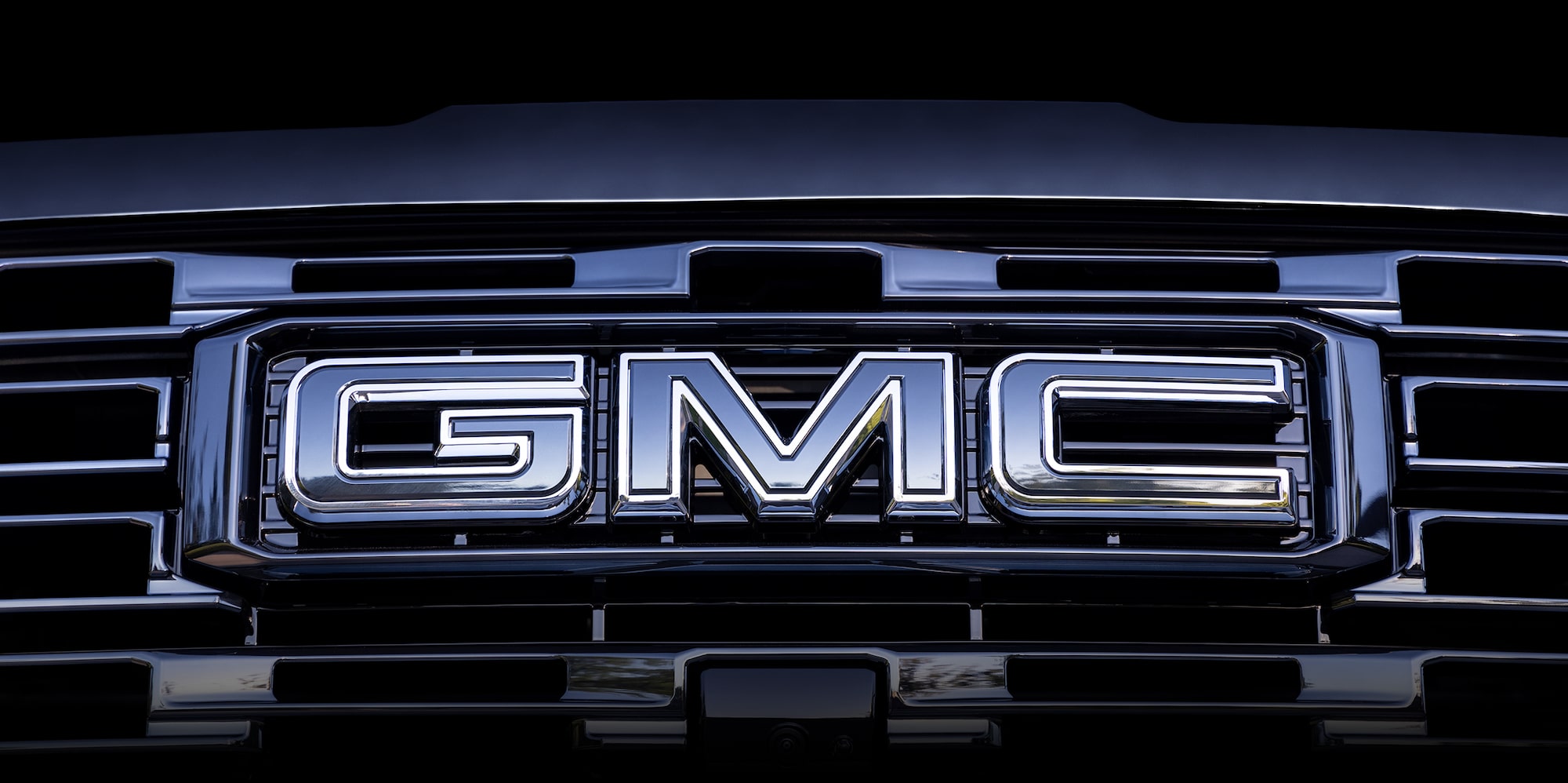 A Close-Up of the Chrome GMC Logo On a Dark Grille with Horizontal Bars, Showing a Camera Below the Badge