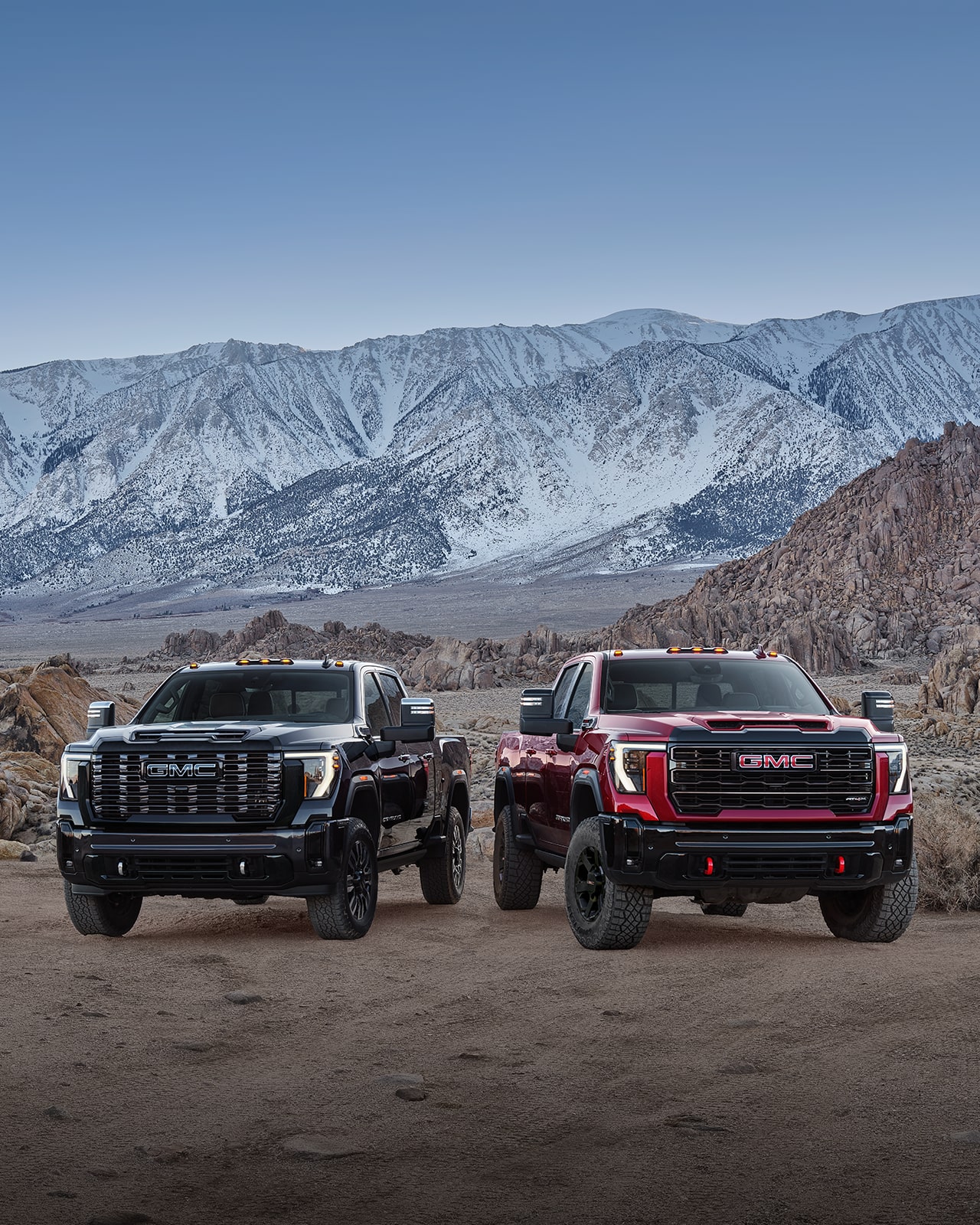 Two 2027 GMC Sierra HD Trucks, One Black and One Red, with Off-Road Tires and Red Recovery Hooks Parked Side By Side On a Rocky Terrain with Snowy Mountains in the Background