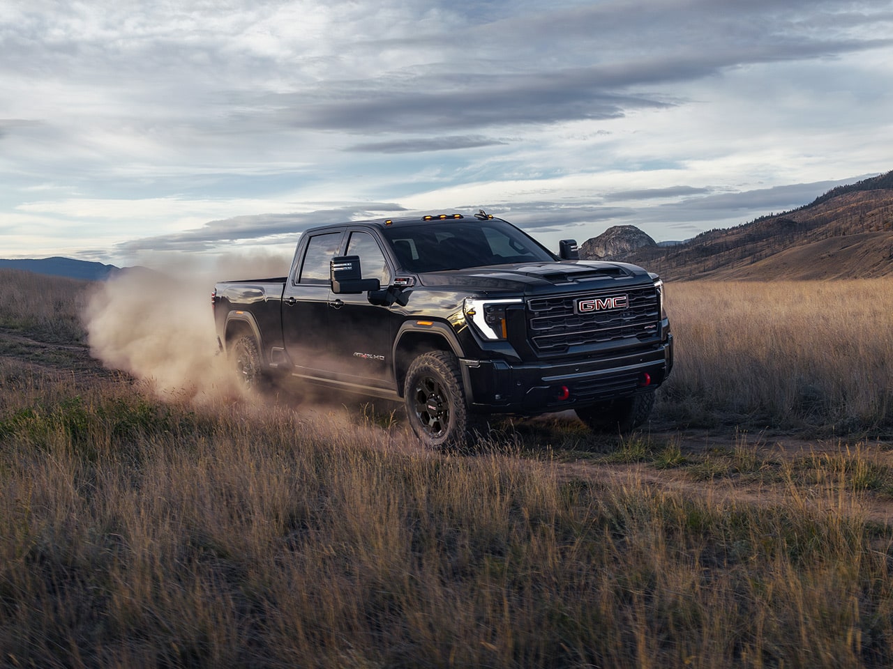 A Black 2027 GMC Sierra HD Truck Kicks Up Dust As It Drives On a Dirt Path Through Dry Grass Fields with Rolling Hills and a Cloudy Sky