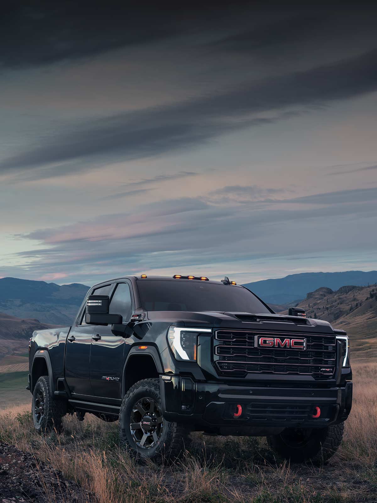 A Dark Gray 2027 GMC Sierra HD AT4X Parked on a Grassy Hill with Mountains under a Cloudy Sky