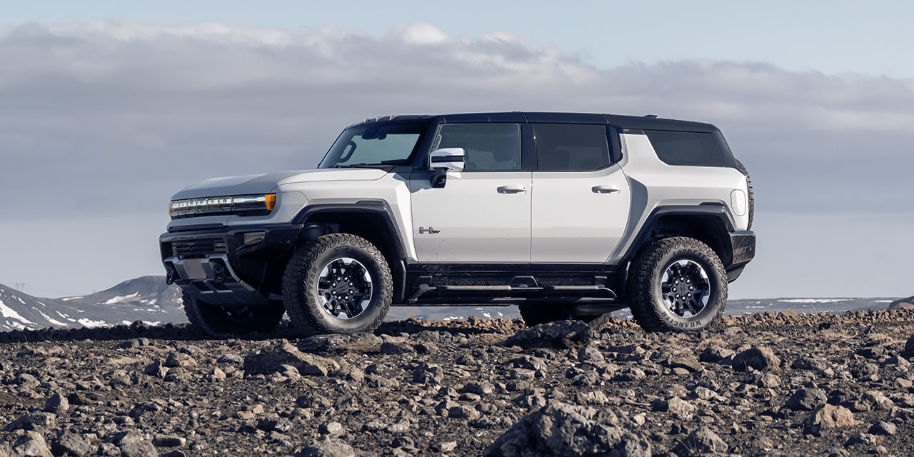 Profile View of a White 2026 GMC HUMMER EV SUV Parked on Rough Terrain with Mountains in the Distance