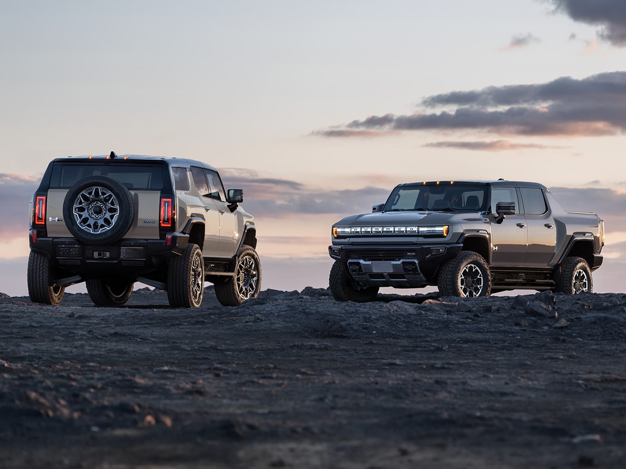 GMC HUMMER EV SUV and HUMMER EV Pickup Truck Parked on Rocky Terrain with a Beautiful Sunset Background
