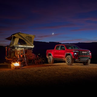 A Person Sits By a Campfire Next To a Red GMC Canyon AT4X Truck with a Rooftop Tent On a Trailer, Under a Twilight Sky with a Crescent Moon