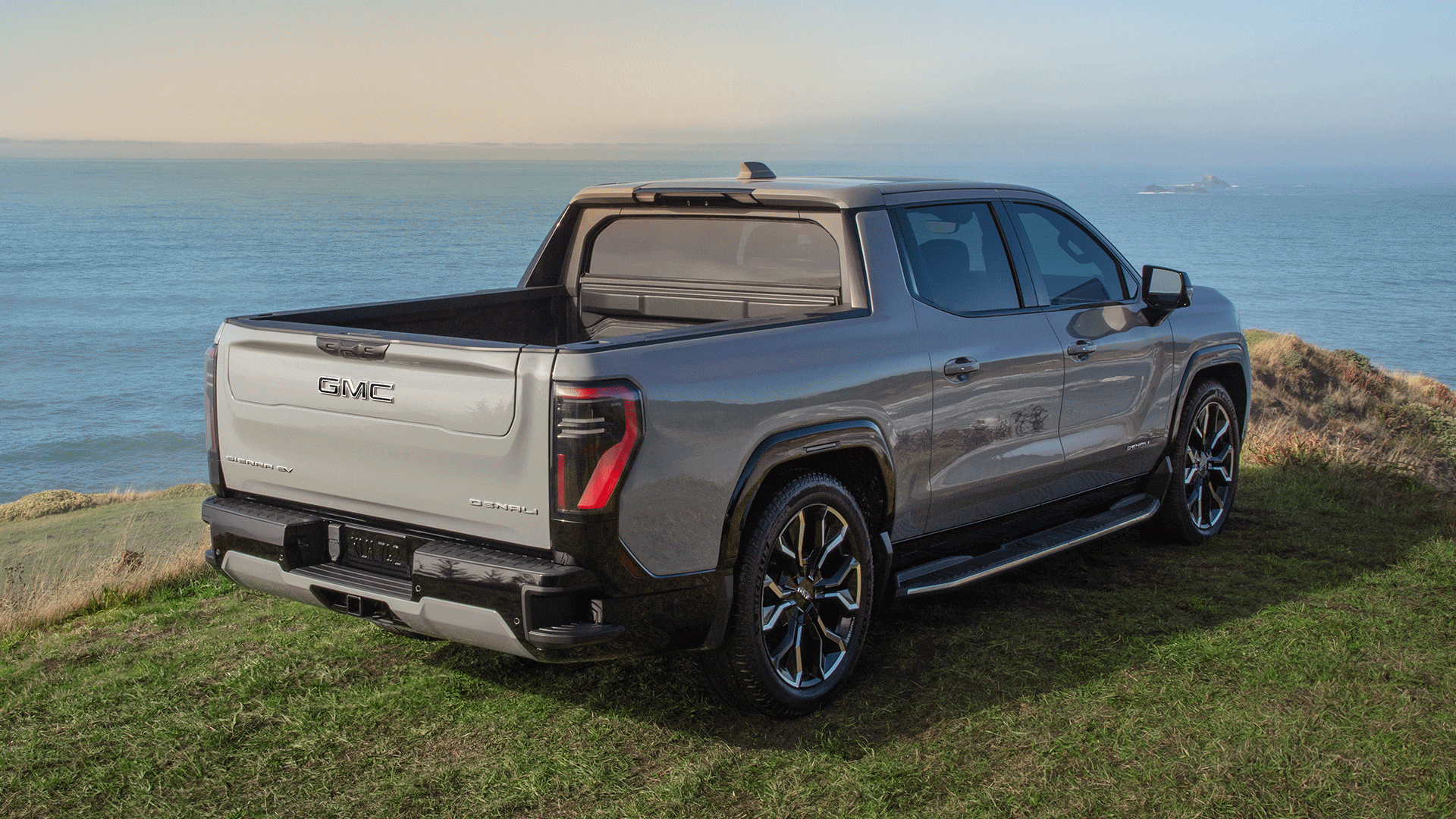 Rear Three-Quarters View of the GMC Sierra EV Parked on a Hill Over Looking a Body of Water with the Truck's Multipro Tailgate Open