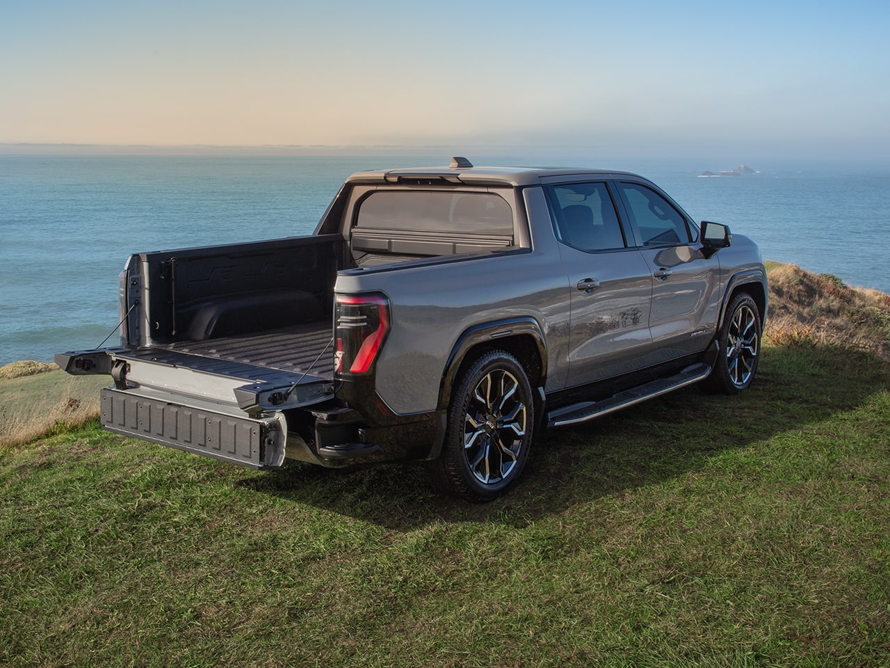 Rear Three-Quarters View of the GMC Sierra EV Parked on a Hill Over Looking a Body of Water with the Truck's Multipro Tailgate Open