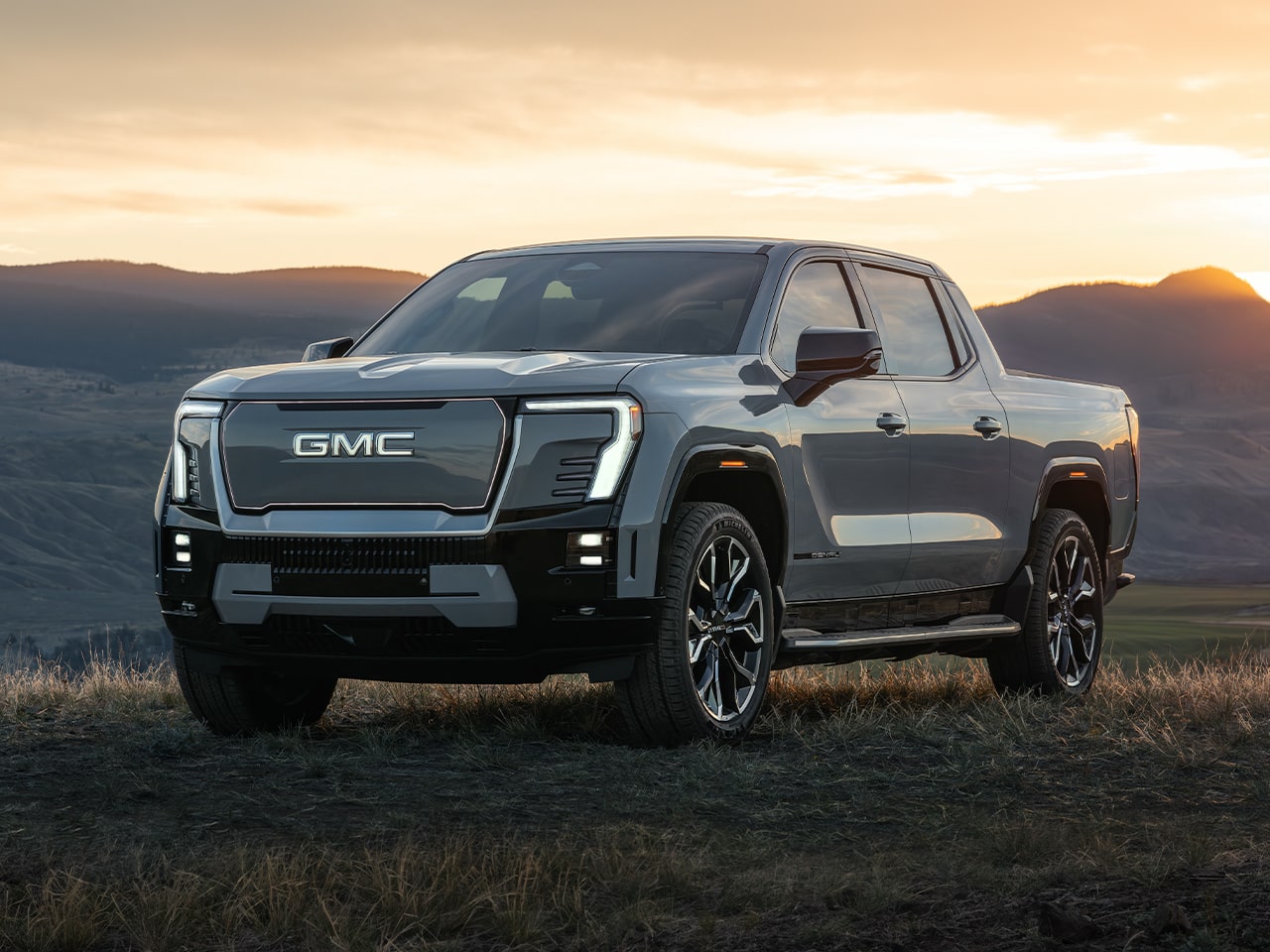Front Three-Quarters View of the GMC Sierra EV Pickup Truck Parked on top of a Grassy Hill at Sunset